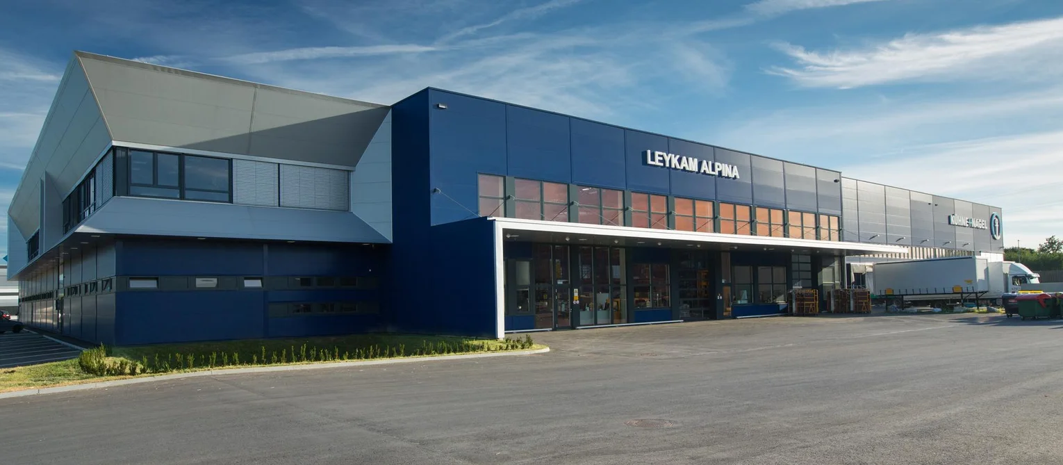 Photo: Leykam Alpina: Exterior of light-grey warehouse and production hall with dark-blue accents. The office area is adjoined at the left of the building.
