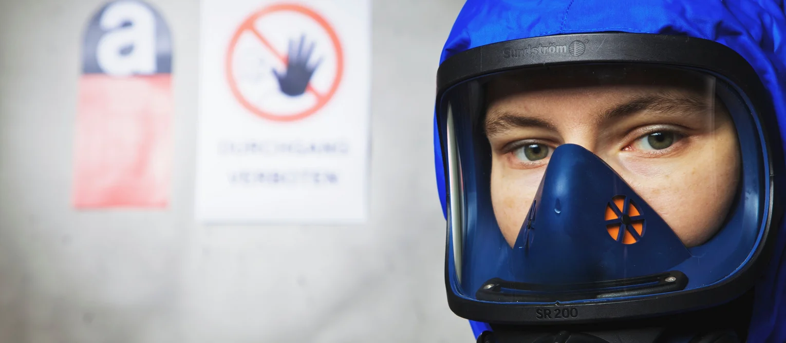 Foto: Asbest-Sanierung: Ein Mann in einem Vollkörperanzug mit Atemschutzmaske; im Hintergrund eine Wand mit Warntafeln