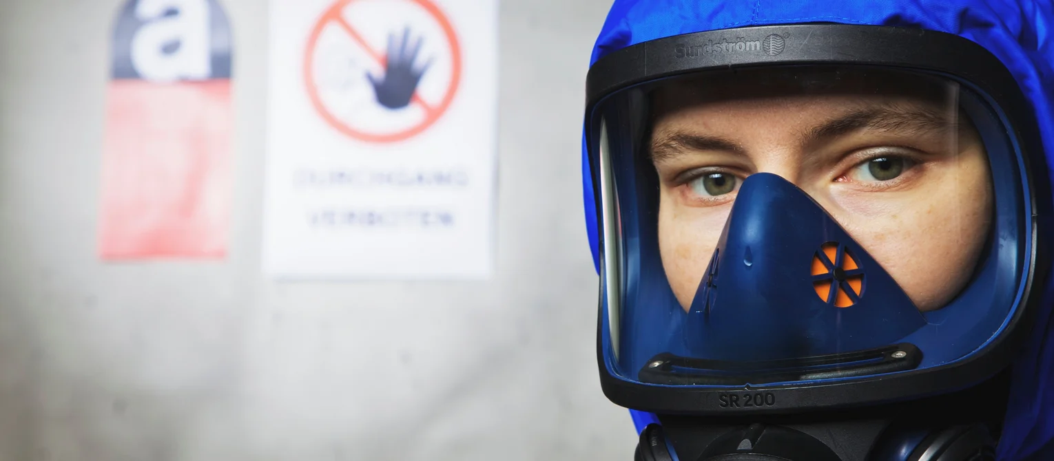 Photo: Asbestos rehabilitation: A man in a full body suit with respiratory mask; in the background a wall with warning signs