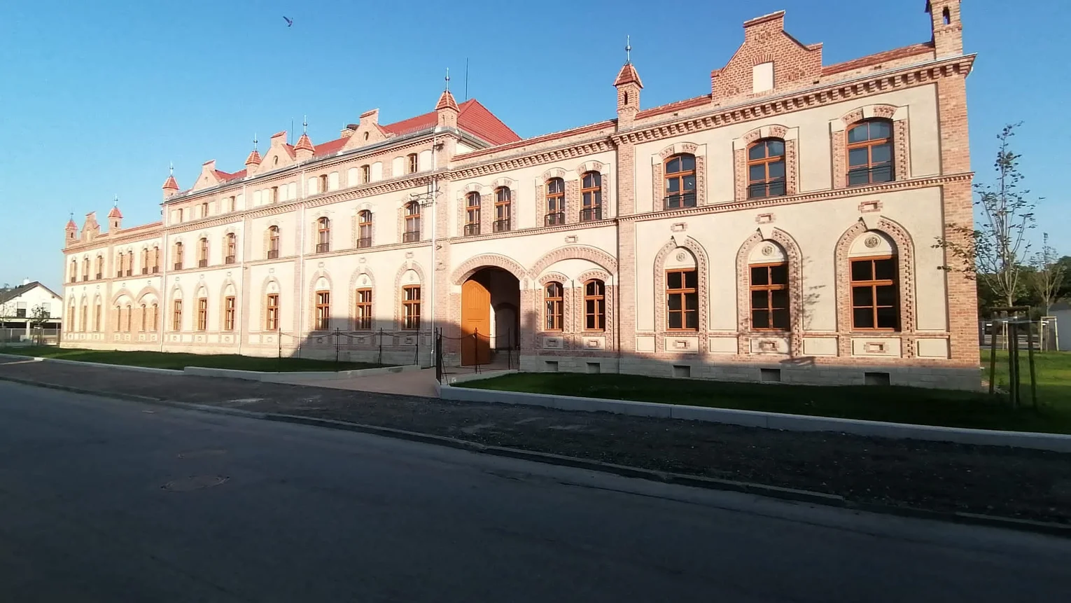 Blick auf die Fassade eines zweigeschossigen historischen Industriebaus mit Backstein-Elementen, einem großen Einfahrtstor, zahlreichen großen Fenstern und kleinen Türmchen an der Dachkante des Gebäudes. Im Vordergrund ein gepflegter Grünstreifen zwischen Gehsteig und Gebäude, davor eine Straße.