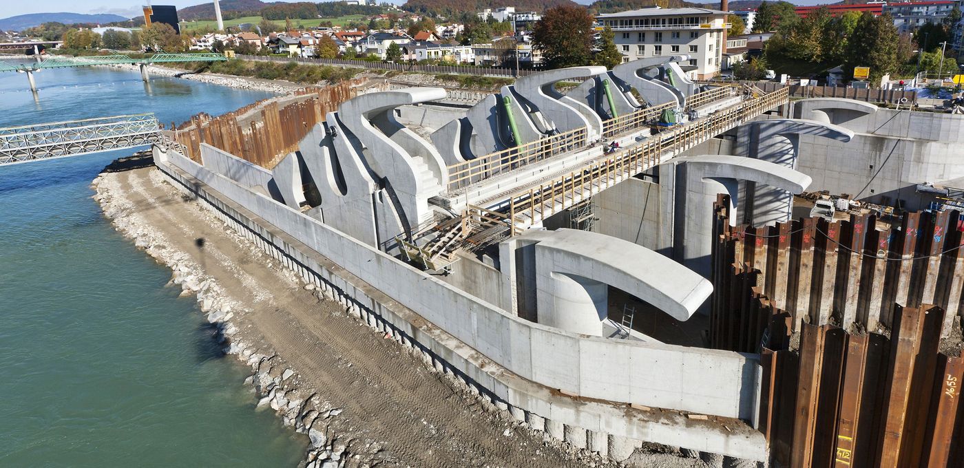Foto: Kraftwerk Lehen: seitlicher Blick auf die in Bau befindlichen Wehranlagen auf der rechten Salzachseite, stromabwärts gesehen; links vorn im Bild die Salzach