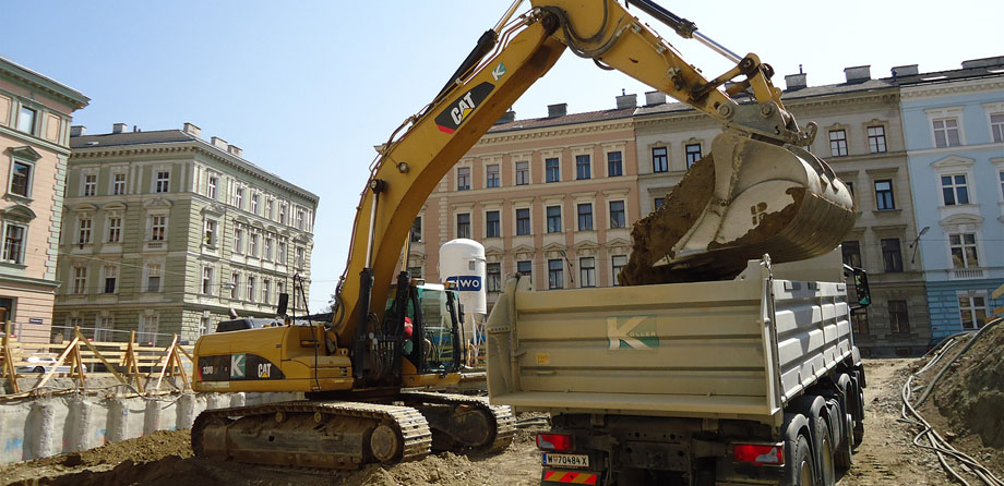 Foto: Ein Raupenbagger belädt einen Lkw mit Erdreich auf einer Baustelle in mitten historischer Gebäude.